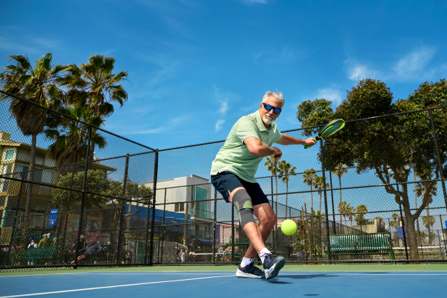 man with white hair, sunglasses, and a green shirt swinging to hit a tennis ball at tennis courts